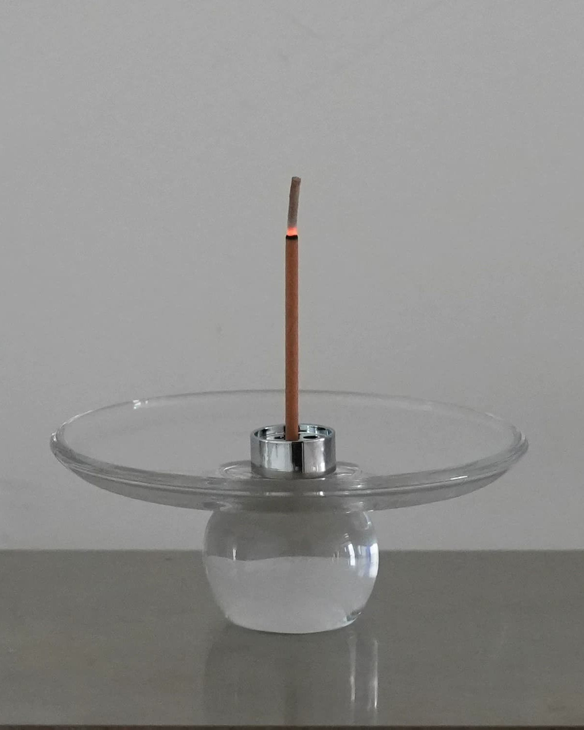 Clear Hour glass incense stick holder with a lit stick standing in one of the nine holes of the steel insert, ash falling into the wide glass dish