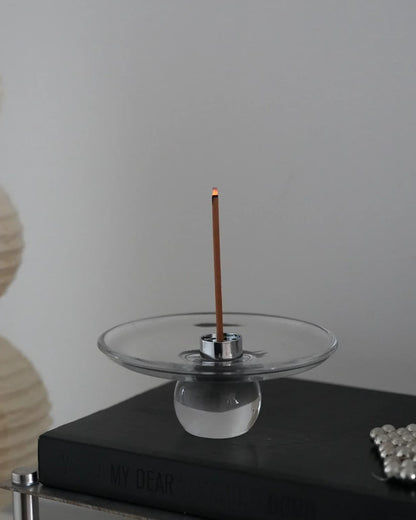 Clear glass incense stick holder on a dark tabletop, a lit stick burning in the removable steel insert