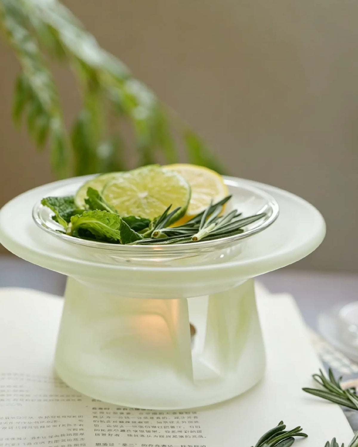 Celadon green glass incense warmer on an open book, citrus slices and rosemary in the dish above a lit tealight