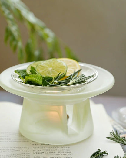 Celadon green glass incense warmer on an open book, citrus slices and rosemary in the dish above a lit tealight