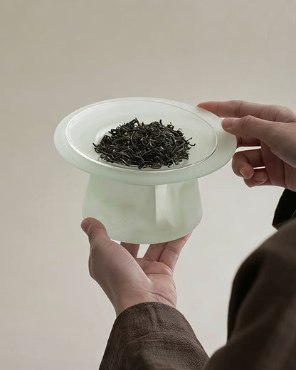 Hands holding a celadon green liuli glass incense warmer with dried tea leaves in the dish