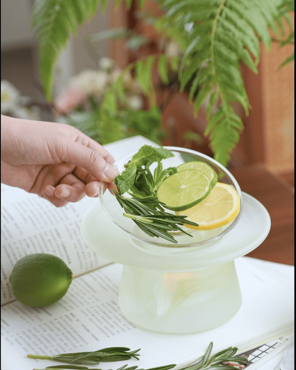 A hand placing rosemary and citrus into the upper dish of a celadon green glass incense warmer