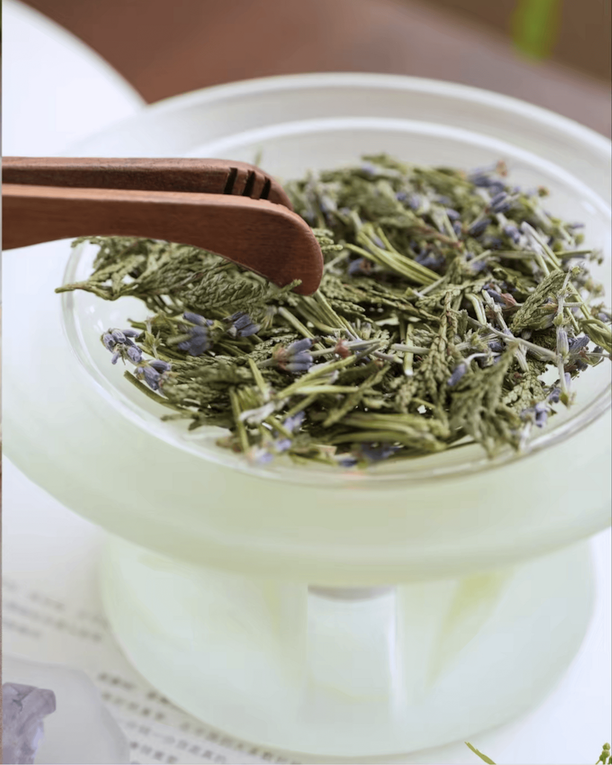 Wooden tongs arranging dried lavender and herbs in the dish of a celadon green glass incense warmer