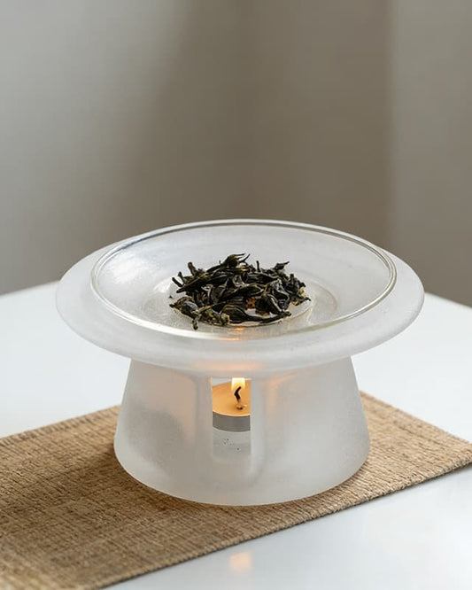 Bone white liuli glass incense warmer on a woven mat, dried tea leaves in the dish and a tealight lit below